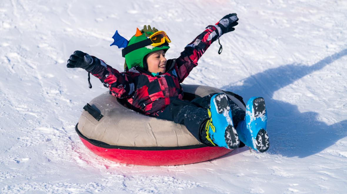Boy wearing winter hat and jacket with outstretched arms riding in a tube tubing on snowy slope