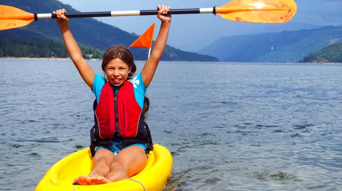 A girl in a yellow sit-upon kayak holds her paddle over her head in Shuswap Lake, British Columbia, among great lake vacations for summer for Seattle-area families