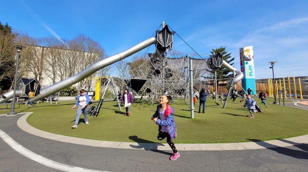 A girl runs in front of the high climber and long tube slide that are part of the Artists at Play playground at Seattle Center among risky adventure playgrounds for thrill-seeking kids