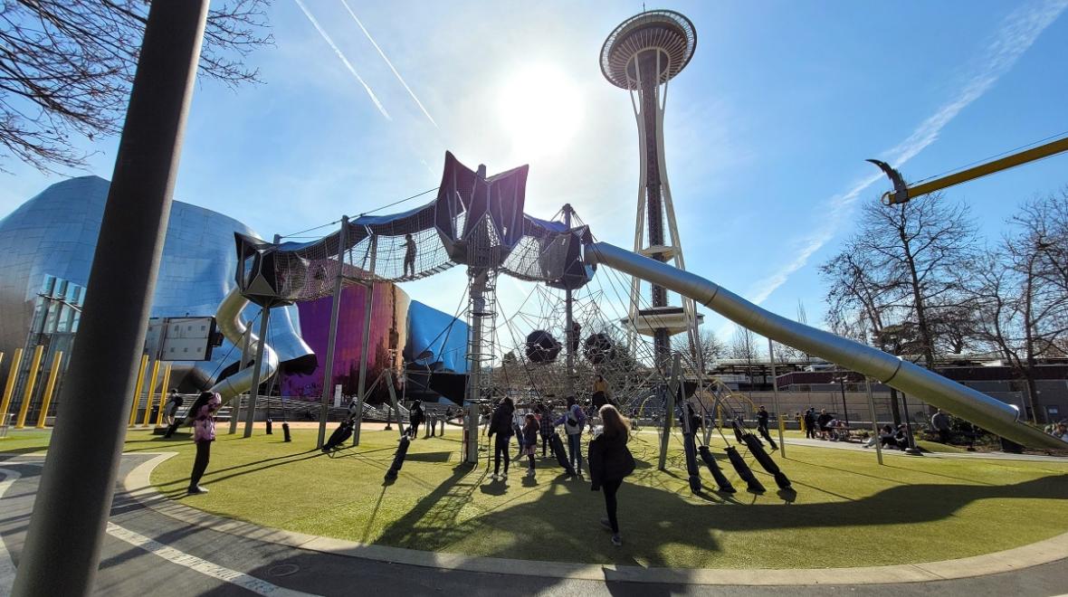 Kids play on the Artists at Play playground at Seattle Center on a sunny spring day. The Space Needle and the Museum of Pop Culture (MoPop) are visible behind
