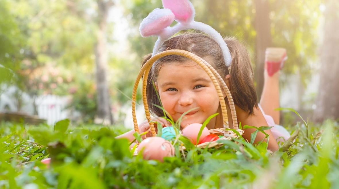 A cute young girl peers through the rounded handle of her Easter basket with colored eggs in it. She is lying in the grass and wearing bunny ears. Best Seattle Easter events for kids and families 2023