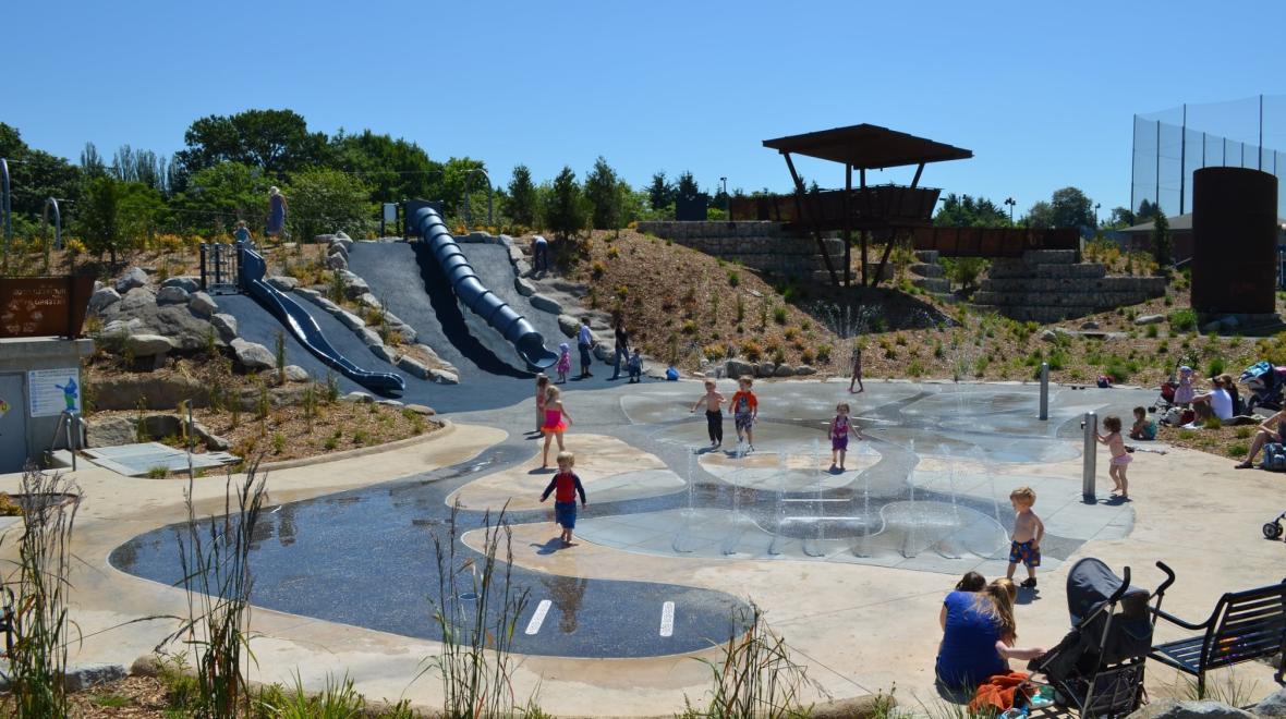 Kids play on the summer spray ground at Seattle's Jefferson Park with two hillside slides in the background, Jefferson Park's playground is among Seattle's most adventurous and fun