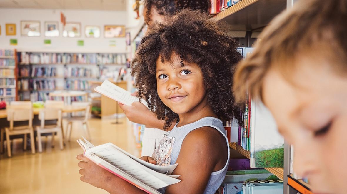 Young girl leaning against a shelf of books holding an open book and smiling at the camera