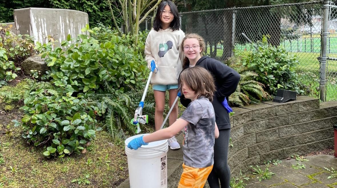 Three elementary kids holding buckets cleaning up an outdoor area