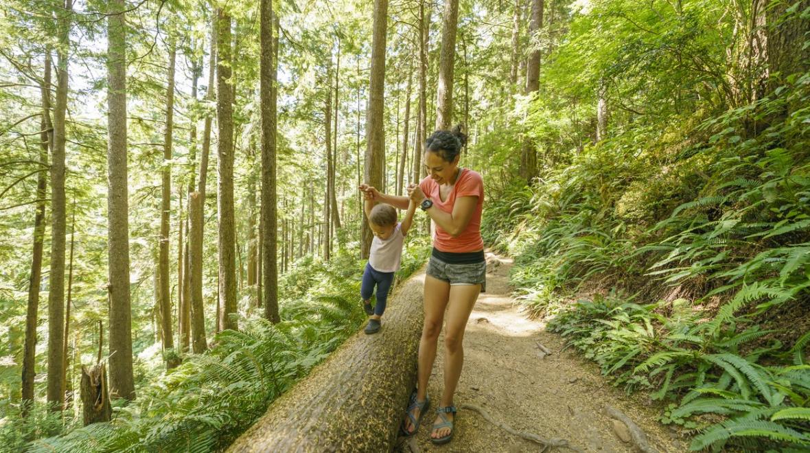 A mom dressed in running clothes stands along a wooded trail. She is holding the hands of her young toddler who is walking along a large fallen tree as sunlight filters through the woods
