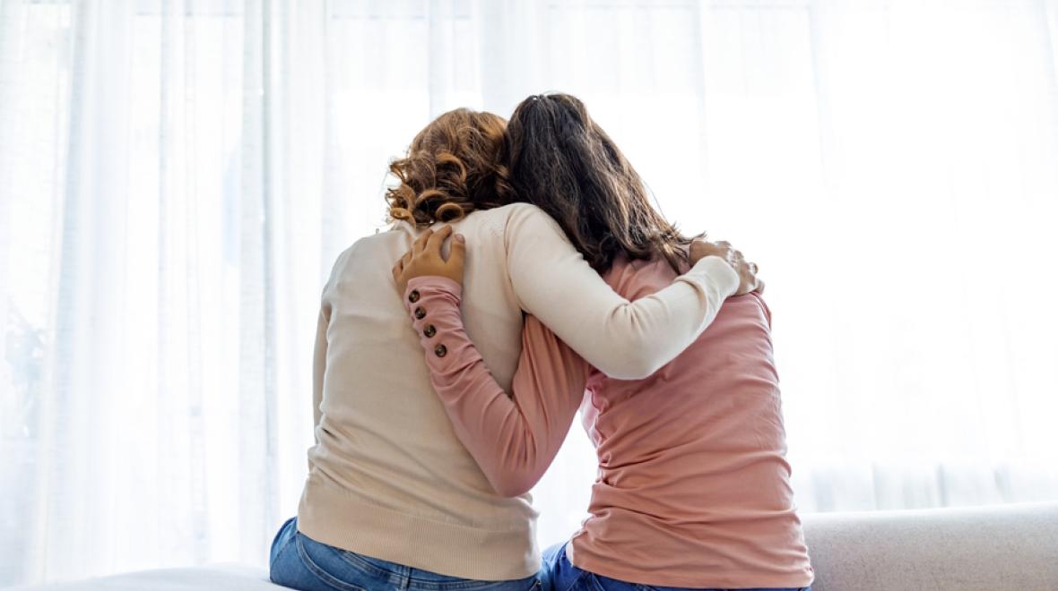 Mom and daughter sitting on a bed with their arms around each other, seen from the back
