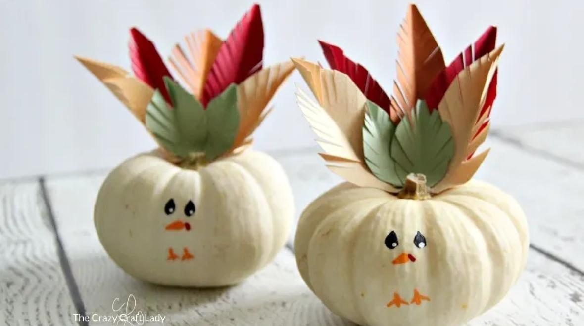 Two small white pumpkins with turkey faces painted on and construction paper leaves at the top