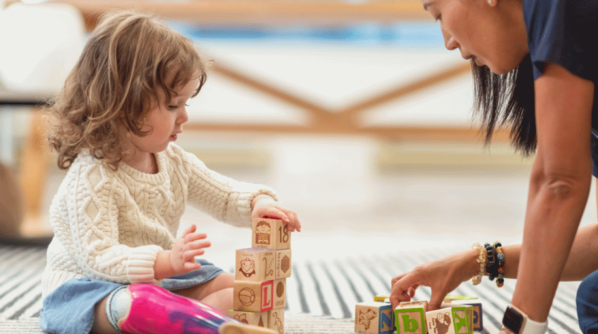 a young girl plays with wooden blocks along with an adult on a rug, safe toys