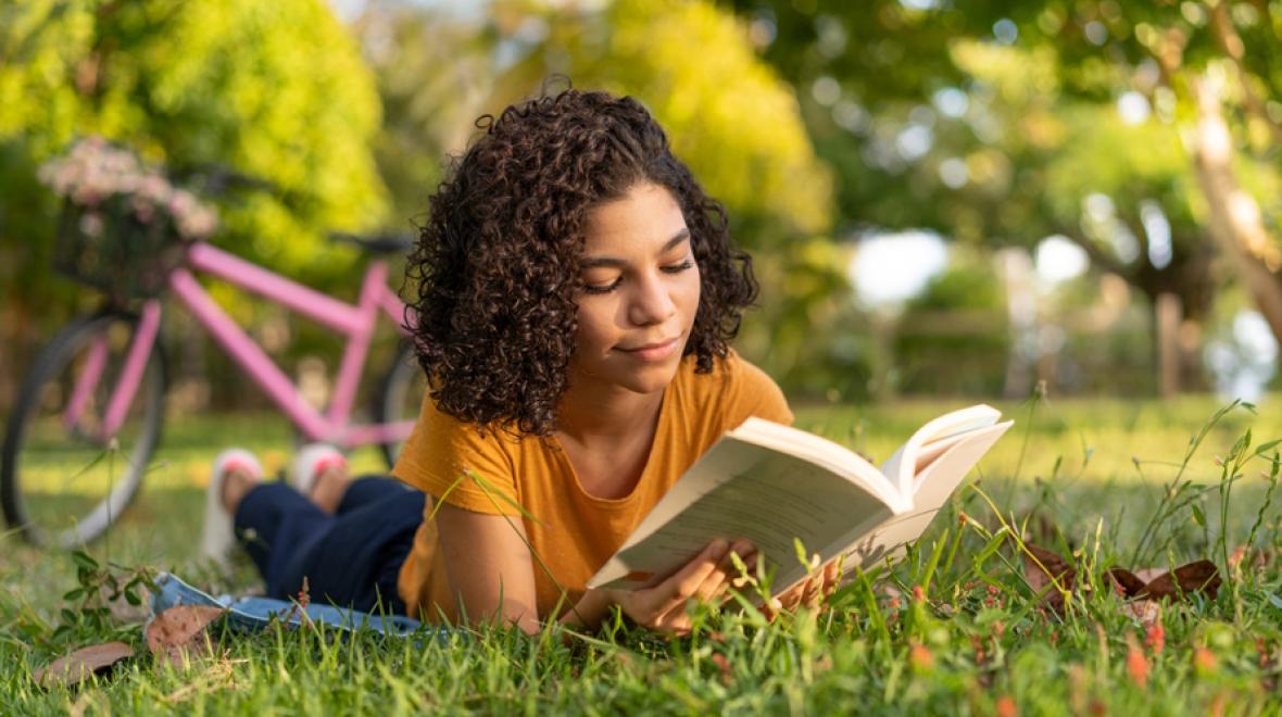 Teenage girl lying in the grass reading a young adult book 