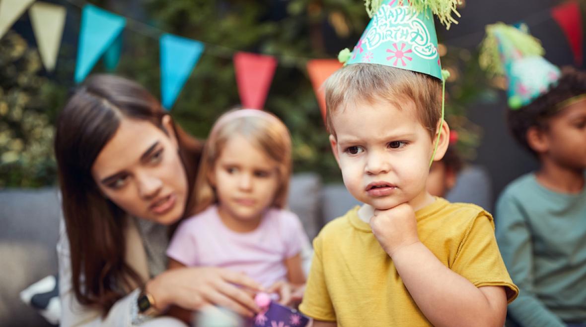 boy looks sad wearing a birthday party hat with a woman and children in back