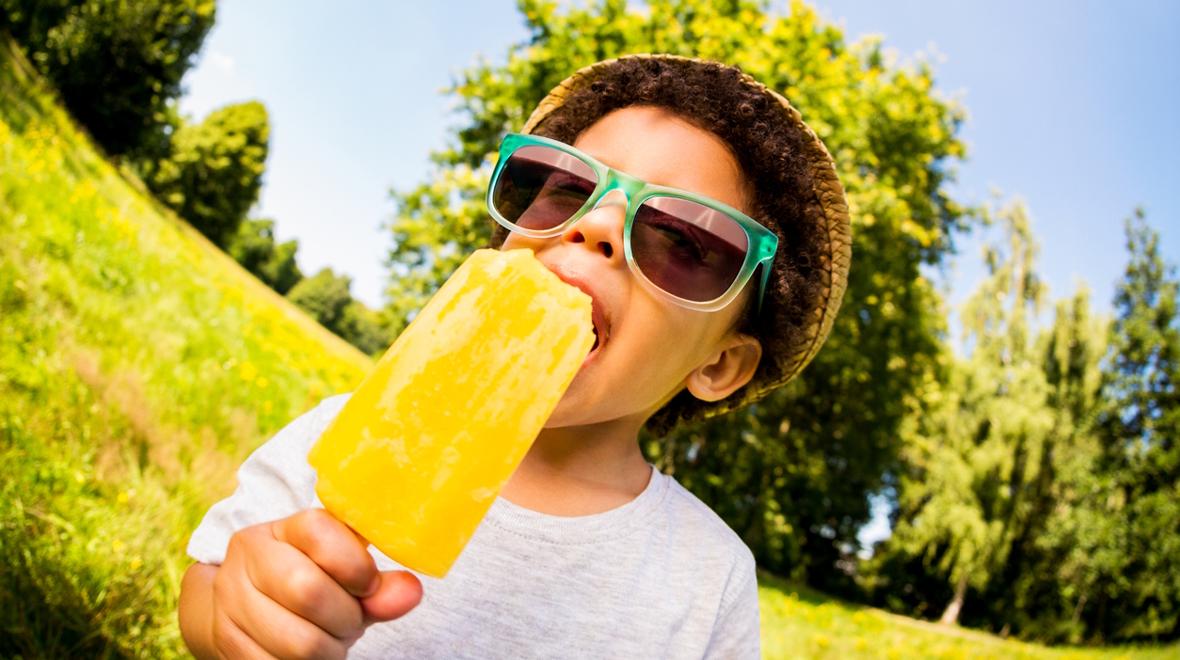 Boy wearing a hat and sunglasses eating a sweet treat in the summer