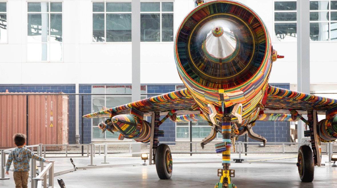young boy walking underneath the wing of a fighter jet decorated with glass beads, part of “The MiG-21 Project” exhibit this weekend at The Museum of Flight