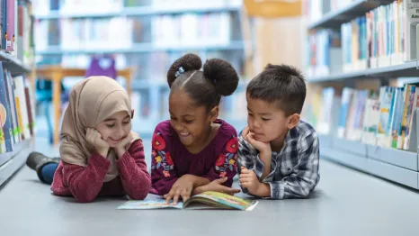 Three kids lying on their stomachs in a library looking at a book together