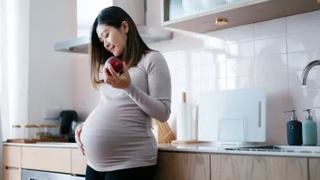 Pregnant woman standing in a kitchen holding an apple and looking down at her belly 