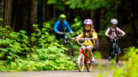 A young girl rides a bike on a bike path with her older sibling and parent following behind. They are wearing helmets and the paved path is lined with green plants indicating summer.