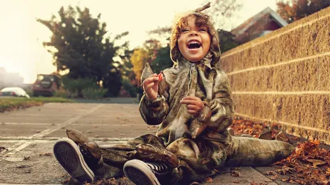 Little boy sitting on the sidewalk smiling wearing a dinosaur Halloween costume