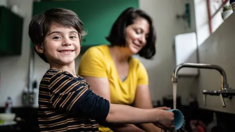Boy washing a mug and smiling at the camera with mom in the background