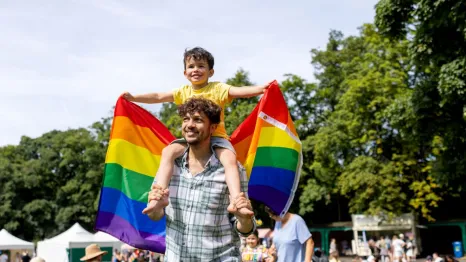 A young boy holds a Pride rainbow flag while on his father's shoulders to celebrate Pride.
