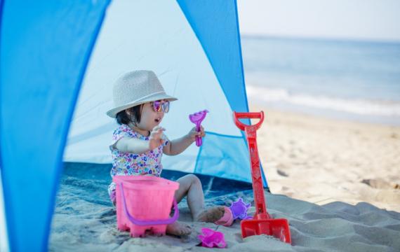 Kid-wearing-sunhat-sitting-in-tent-by-the-beach, sun protection for kids