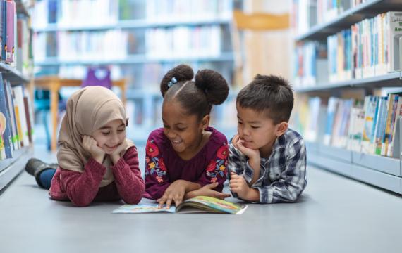 Three kids lying on their stomachs in a library looking at a book together