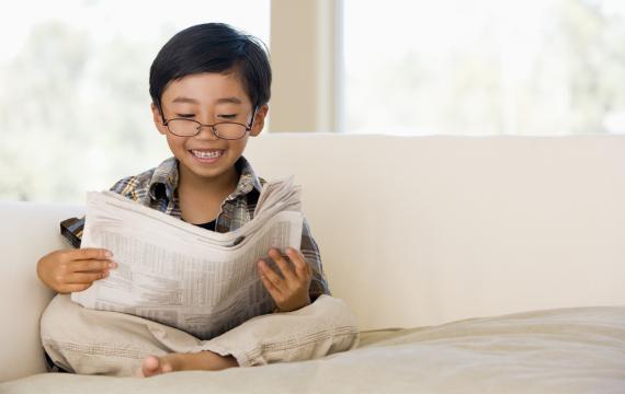 Young boy wearing glasses sitting on a couch reading a newspaper