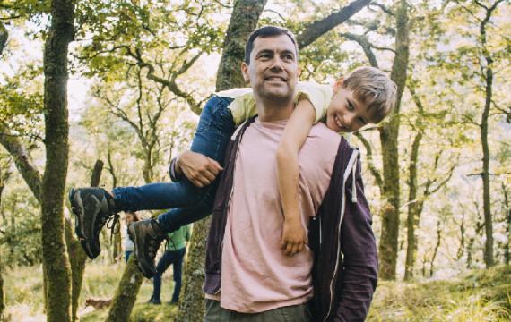 Little boy is smiling at the camera as he is being fireman carried by his father through the woodlands.