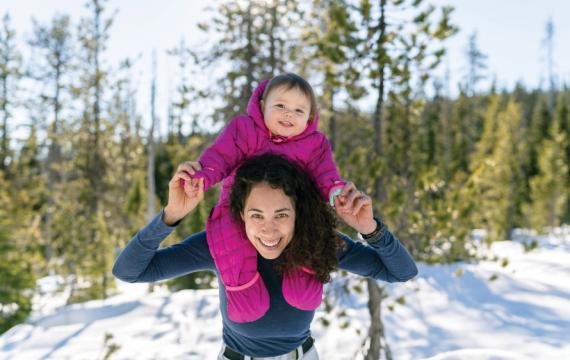 Woman holding a smiling baby on her shoulders in the woods with snow on the ground