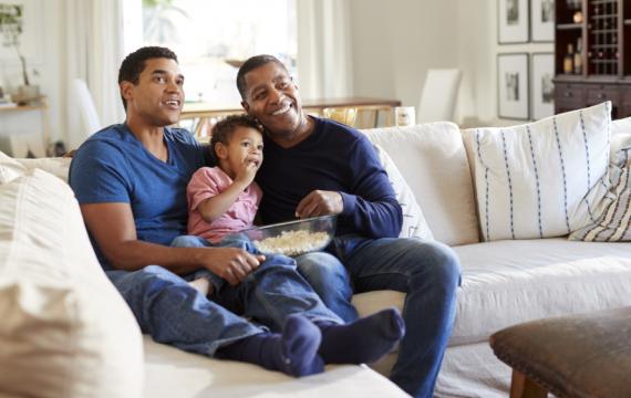 Grandpa, dad and son watching tv on a couch eating popcorn