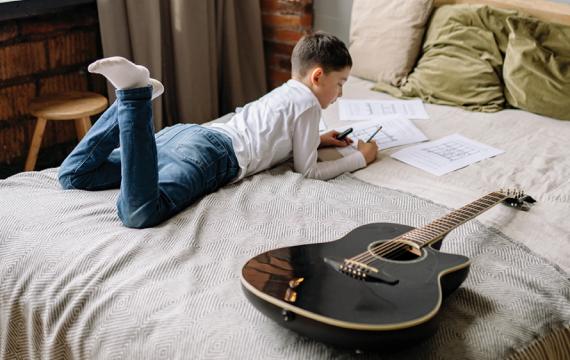 Young boy lying on his bed next to a guitar writing on a paper