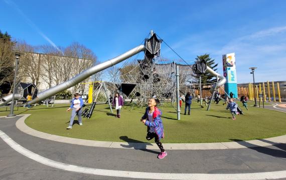 A girls runs in front of the tall climbing play structure at the Artists at Play playground at Seattle Center, among the best most adventurous playgrounds in Seattle and beyond