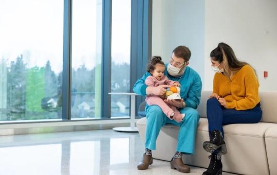 Young baby girl sitting in a doctor's lap with mom sitting next to them on a couch