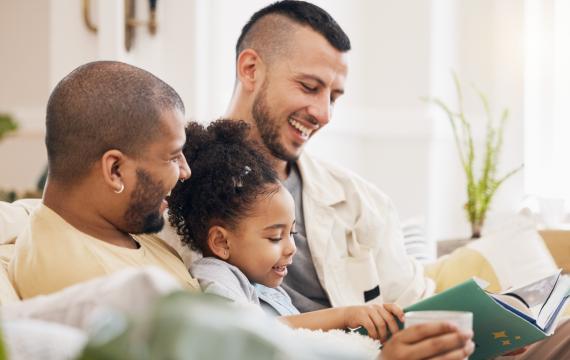 Two dads sitting on the couch with their daughter reading a book with LGBTQ characters