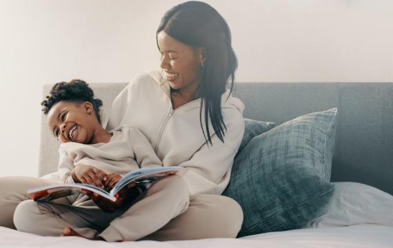 Preschool age girl laughs happily while sitting with her mom reading a story book. 