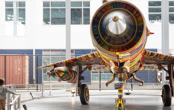 young boy walking underneath the wing of a fighter jet decorated with glass beads, part of “The MiG-21 Project” exhibit this weekend at The Museum of Flight
