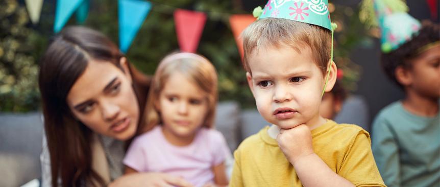 Little boy scowling in a party hat. Woman and little girl look on.