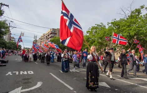 People in Norwegian traditional costumes walk in Ballard Syttende Mai parade holding Norwegian flags