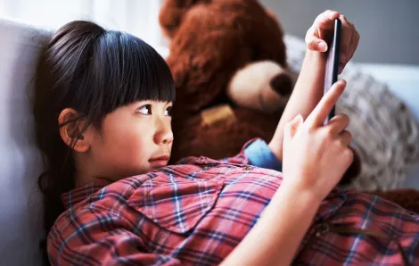 Girl at home looking at tablet sitting on couch with teddy bear beside her