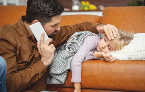 dad holding a hand to his sick daughter's forehead while holding the phone