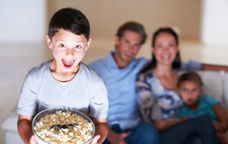 boy holding a bowl of popcorn with a plastic spider in it watching a kids Halloween movie with his parents in the background