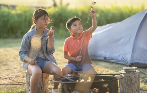 Two young siblings ages 9 and 6 roasting marshmallows by a campfire at a campsite with their tent in the background
