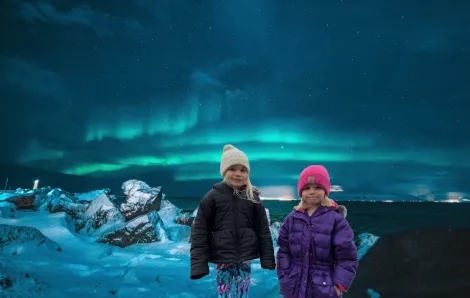 Two girls in winter coats and hats shown in front of a background of the Northern Lights seen in Iceland in this photoshopped virtual travel image
