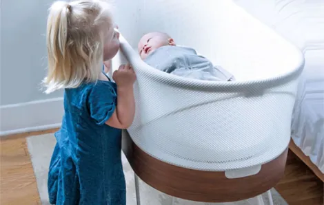 Little girl peeking over looking at sleeping baby in a SNOO bassinet