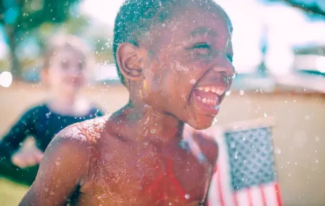closeup of a boy running through a sprinkler with a flag and other kids in the background