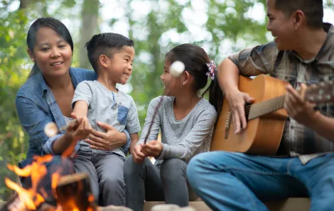 family of four roasting marshmallows over a fire while dad plays a guitar