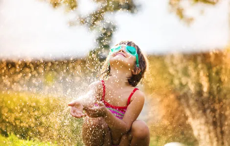 little girl wearing goggles looking skyward while sitting in the stream of a backlit sprinkler
