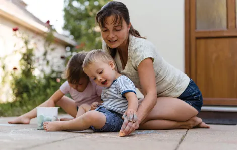 mother sitting on the ground outside her house playing with sidewalk chalk with her two children