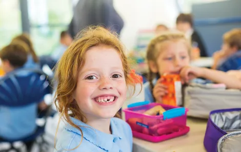 smiling redheaded girl sitting at a lunch table at school with lunchboxes and kids in the background