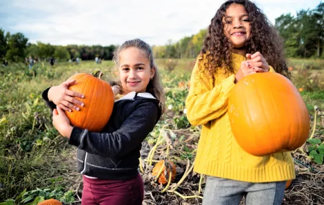 Two elementary-age girls looking at the camera and holding their pumpkin choices at a pumpkin patch in fall best pumpkin patches around Tacoma, South Sound