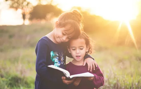 two sisters standing in a field holding a book with the sun behind them
