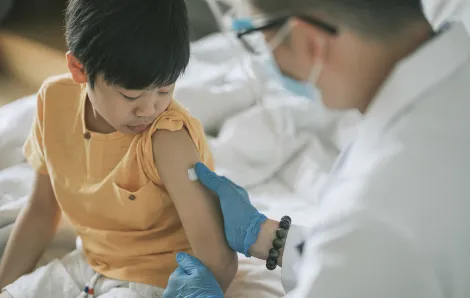 small boy looking down at his arm as a masked doctor puts a bandaid over a vaccine shot poke